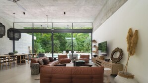 A modern living room with a large window, a brown sofa, a dining area, and a TV on the wall. - Casa Taman, Beachside villa by Elite Beach Villas (Cobano)