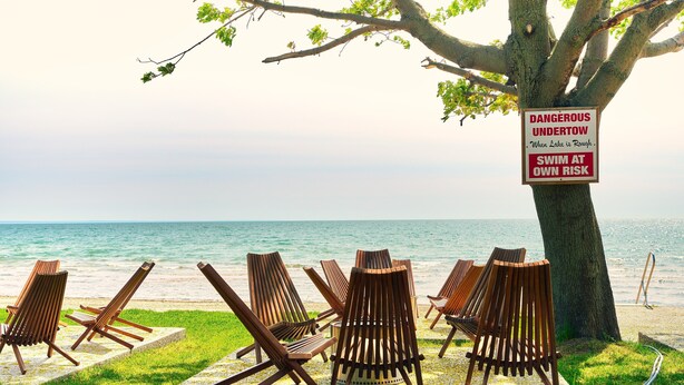 A beachside setting with wooden lounge chairs arranged under a tree, a sign warning of dangerous undertow, and a view of the ocean. - Suite 206: Beautiful 2-Bedroom Beachfront Suite (Wainfleet)