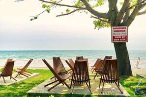 A beachside setting with wooden lounge chairs arranged under a tree, a sign warning of dangerous undertow, and a view of the ocean.