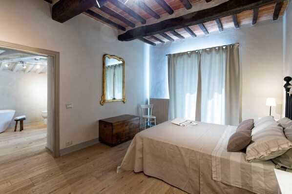 Un dormitorio con suelo de madera, una cama con colcha color crema, una mesita de noche, una silla, un baúl de madera, un espejo y una ventana con cortinas.