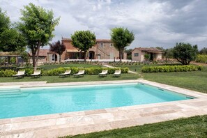 Una piscina con sillas de descanso, rodeada por un césped cuidado y un edificio de piedra con varias ventanas y puertas.