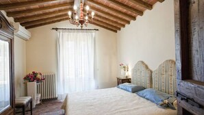Um quarto com uma cama grande, um candelabro, um radiador, uma janela com cortinas e uma pequena mesa com um vaso.
