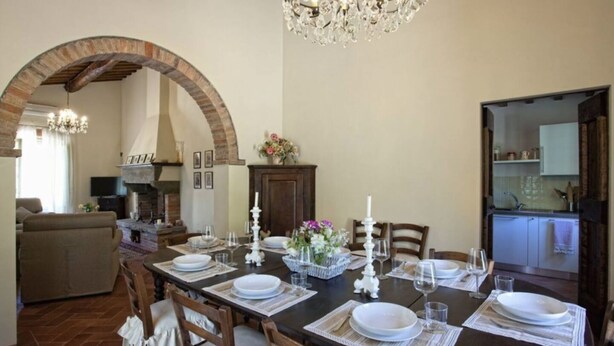 Uma área de refeições com um arco de tijolos, um candelabro e uma cozinha visível ao fundo.