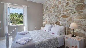Quarto com parede de pedra, cama com roupa de cama branca, mesa de cabeceira com abajur e janela com cortinas.