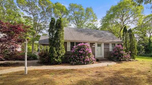 Einbungalow mit Veranda, umgeben von alten Bäumen und blühenden Sträuchern.