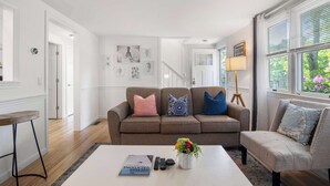 Ein Wohnzimmer mit braunem Sofa, einem weißen Couchtisch und einer Lampe. An der Wand hängen gerahmte Bilder und es gibt ein Fenster mit Jalousien.