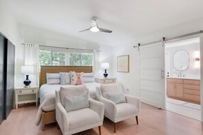 A bedroom with a bed, two armchairs, a nightstand, a bedside table, a ceiling fan, a window with blinds, a door, and a bathroom with a sink and mirror.