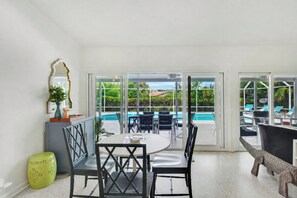 A spacious room with a dining table, chairs, a large mirror, and a view of a pool and greenery outside.