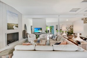 A modern living room with a fireplace, a television, and a dining area with a table and chairs. - Sandpiper Hideaway- Lovely home with pool and just steps away from the beach (Jekyll Island)