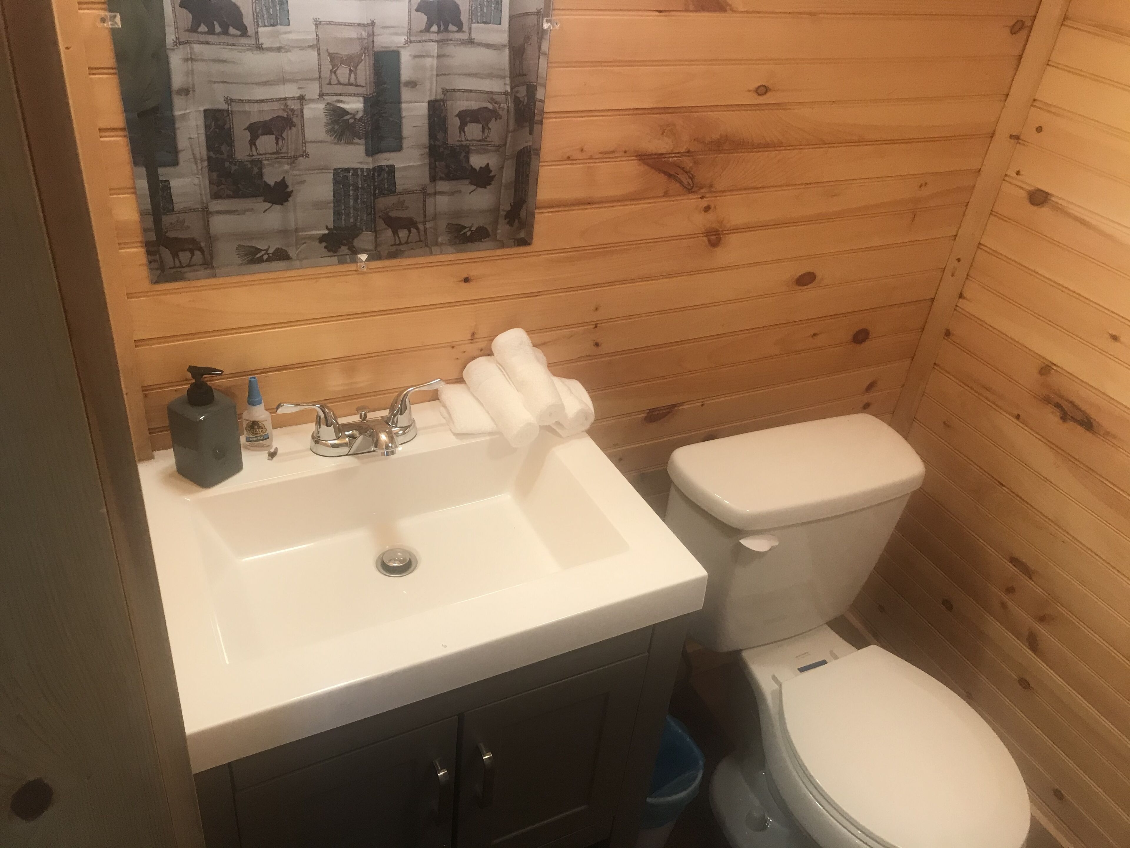 Een badkamer met een wand van houten panelen, een witte wastafel met zeepdispenser, een toilet en een blauwe prullenbak.