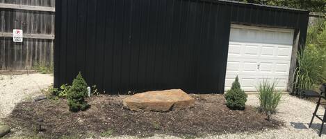 Et sort metalskur med hvid garageport, omgivet af grus, små buske og en stor sten.