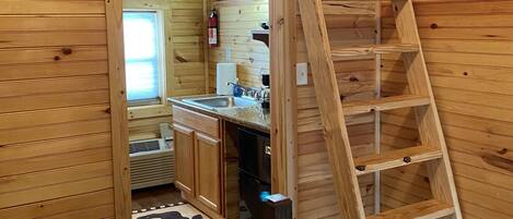 A wooden cabin interior with a loft bedroom, a kitchen area, and a ladder leading to the loft.