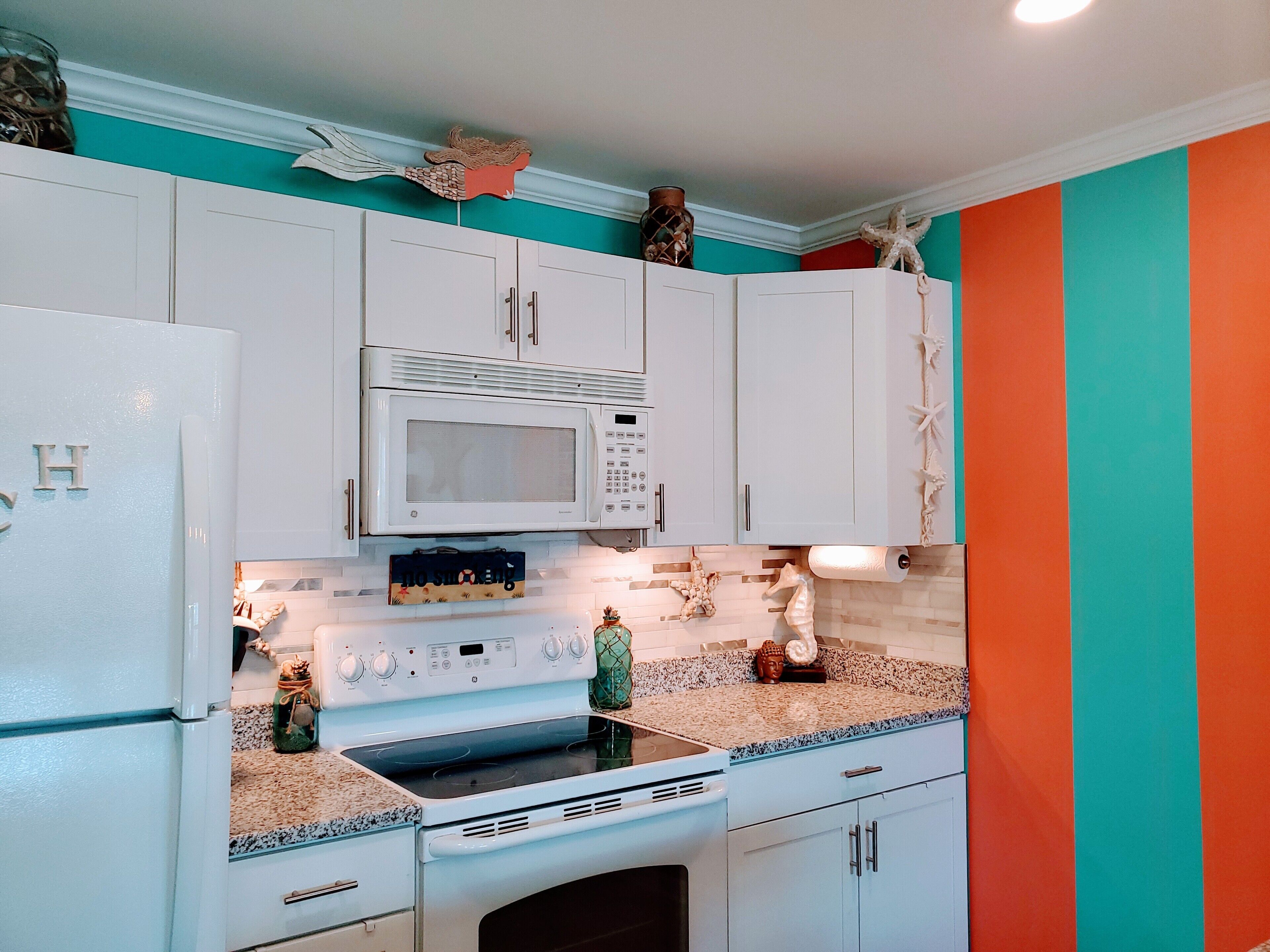 Una cocina con armarios blancos, un microondas y una estufa. Hay elementos decorativos sobre el mostrador y una pared en tonos aguamarina y naranja.