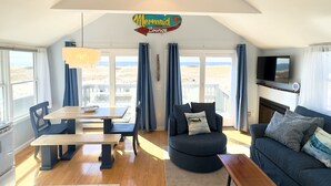 A cozy living room with a wooden table, a dark blue sofa, and a television mounted on the wall. - Beachfront on our private beach!  Siren Cottage - Pipers' Shore (North Truro)