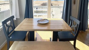 A dining area with a wooden table and chairs, a chandelier, and a view of the beach. - Beachfront on our private beach!  Siren Cottage - Pipers' Shore (North Truro)