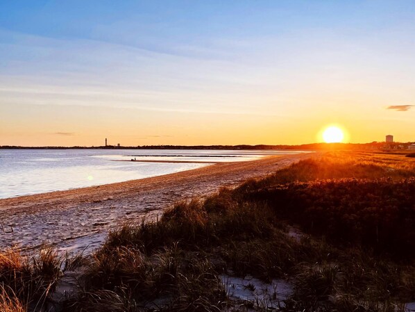 En strand med solnedgang, roligt vand og bygninger i det fjerne.