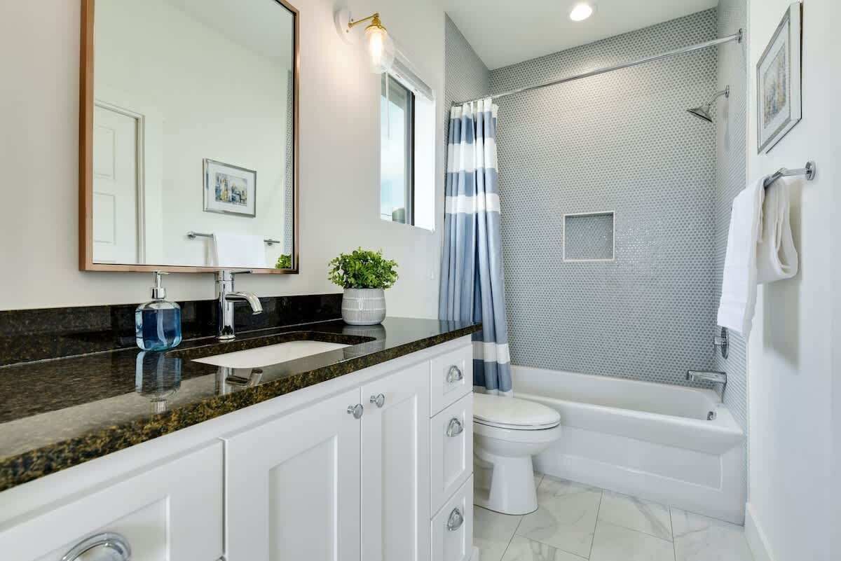 A modern bathroom with a granite countertop, a white sink, a large mirror, a shower with a curtain, and a white toilet.