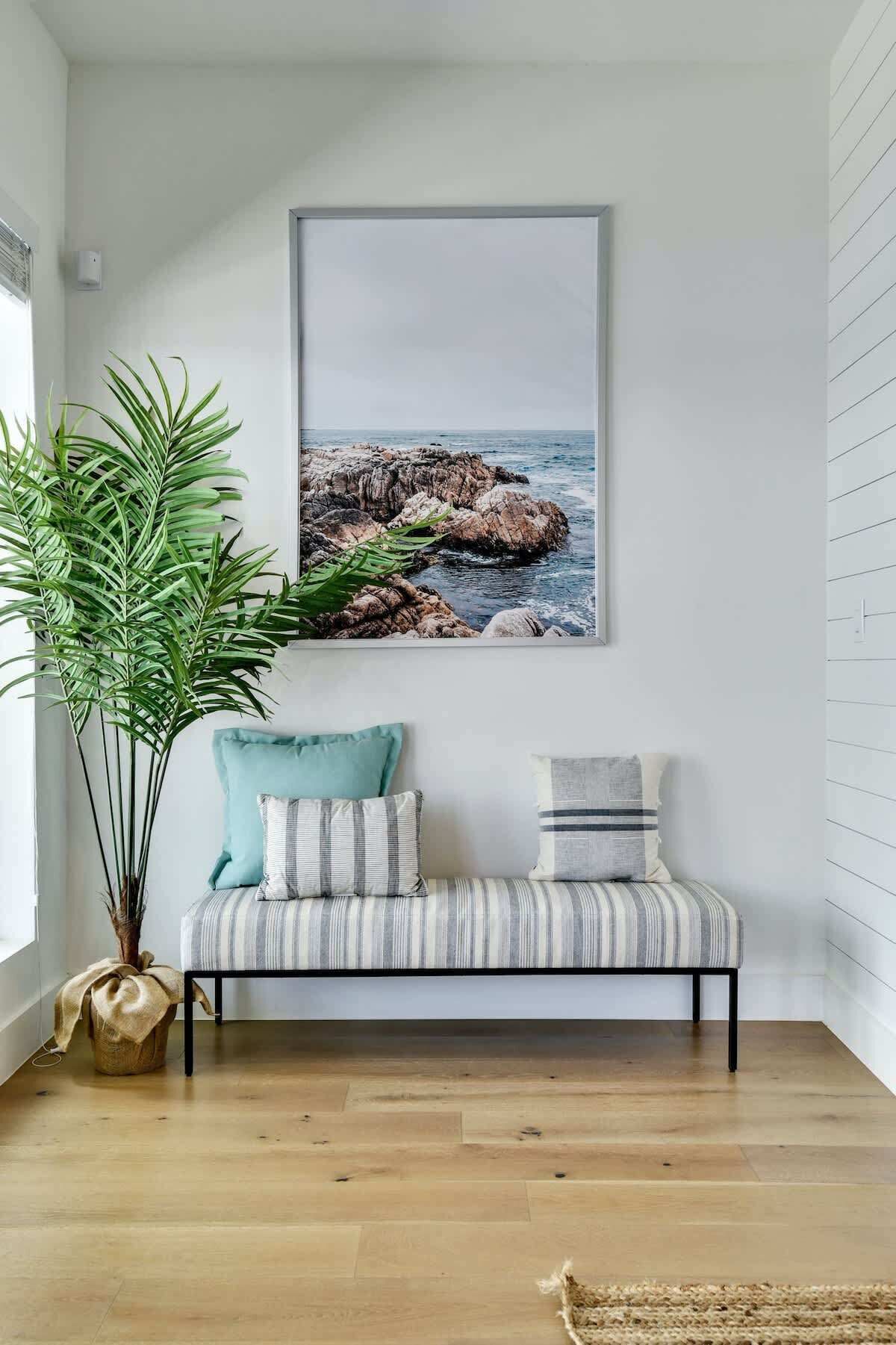 A room with a wooden floor, a bench with cushions, a large potted plant, and a framed picture of a rocky coastline.