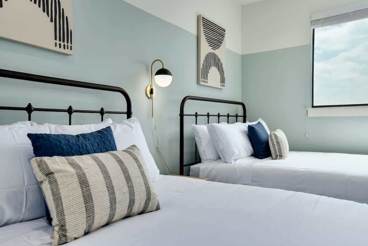 A hotel room with two beds, each with white bedding and multiple pillows. There is a wall-mounted light fixture between the beds.