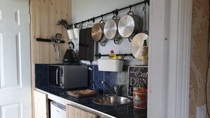 Una cocina compacta con microondas, hervidor y ollas colgantes. Un letrero dice 'Eat Drink Sugar'.