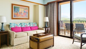 A hotel room with a sofa, a wooden chest, a table with a lamp, and a balcony with chairs.