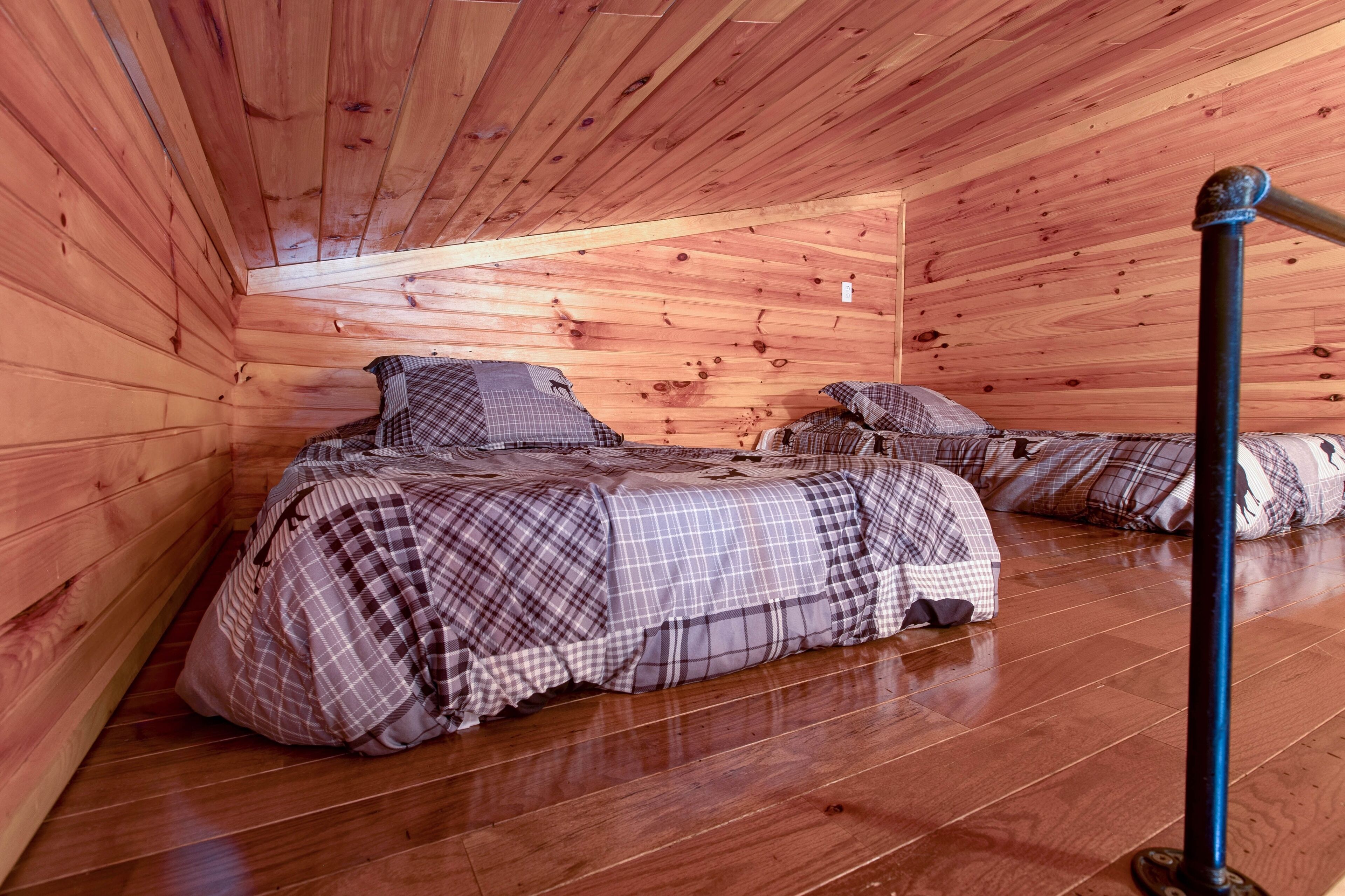 A cozy attic bedroom with wooden walls and a sloped ceiling. There are two beds with plaid bedding and pillows.