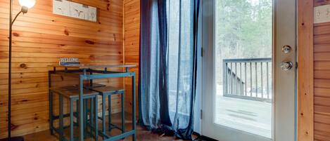 Interior de una cabaña de madera con un letrero de bienvenida, una mesa con sillas, una lámpara de pie y una puerta que da a una terraza.