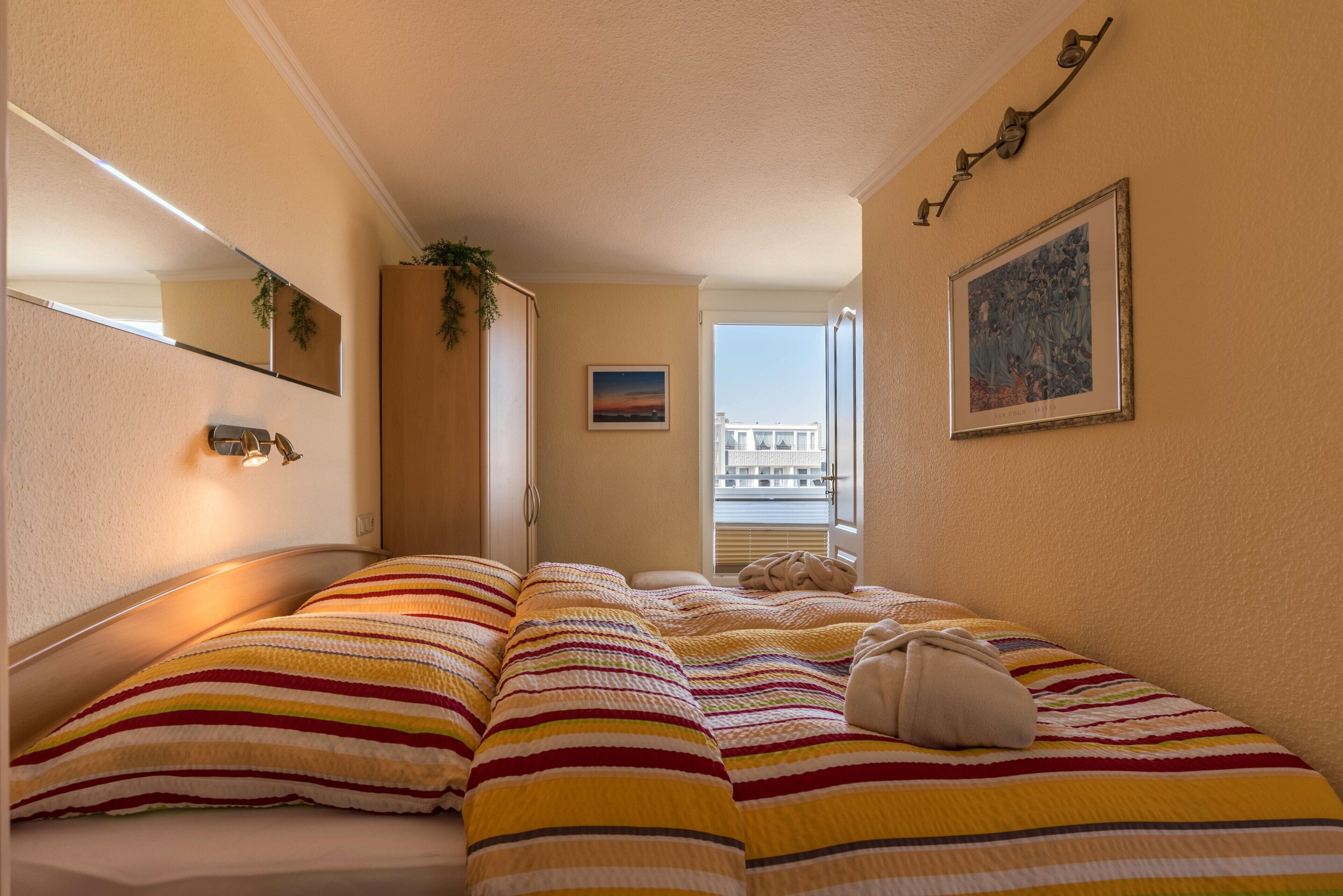 A hotel room with two beds, a window with a view, a painting on the wall, and a plant on the window sill.