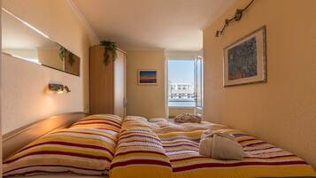 A hotel room with two beds, a window with a view, a painting on the wall, and a plant on the window sill.