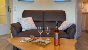 A brown leather sofa with a decorative pillow, two framed pictures on the wall, a bottle of wine, two wine glasses, and a small table with a decorative plate and flowers.
