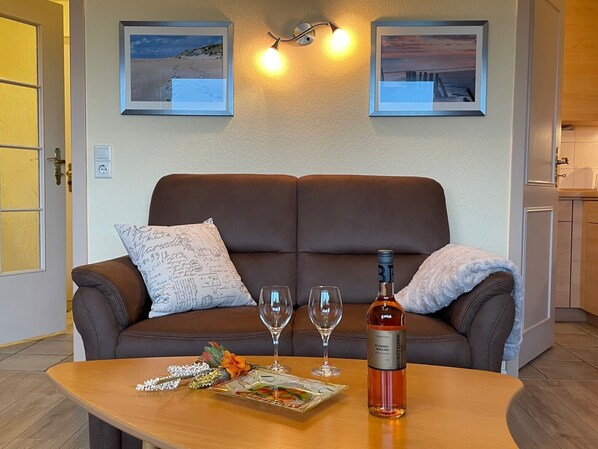 A brown leather sofa with a decorative pillow, two framed pictures on the wall, a bottle of wine, two wine glasses, and a small table with a decorative plate and flowers.
