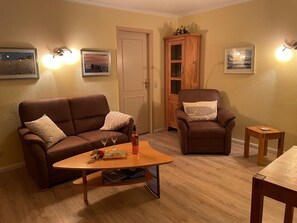 Una sala de estar con un sofá de cuero marrón, una mesa de centro de madera y un sillón marrón. Hay dos cuadros en la pared, un mueble de madera y una puerta.