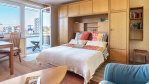 A bedroom with a bed, a wooden wardrobe, a bookshelf, a dining area with a table and chairs, and a view through a glass door.