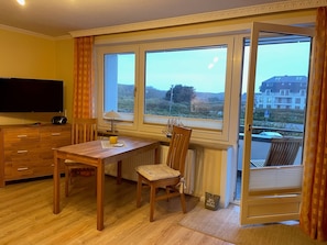 A room with a wooden desk, a chair, a television, and a window with a view of buildings and greenery.