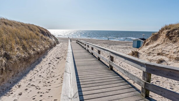 Ein hölzerner Steg führt zu einem Strand mit Dünen und blauem Himmel.