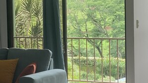 Et værelse med en grå sofa, en gul pude og udsigt til en balkon med grønt.