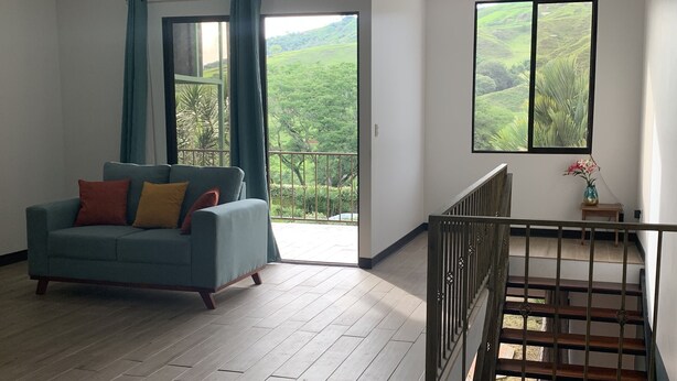 En rummelig stue med en blå sofa, en balkon med udsigt til grønne bakker og en trætrappe.
