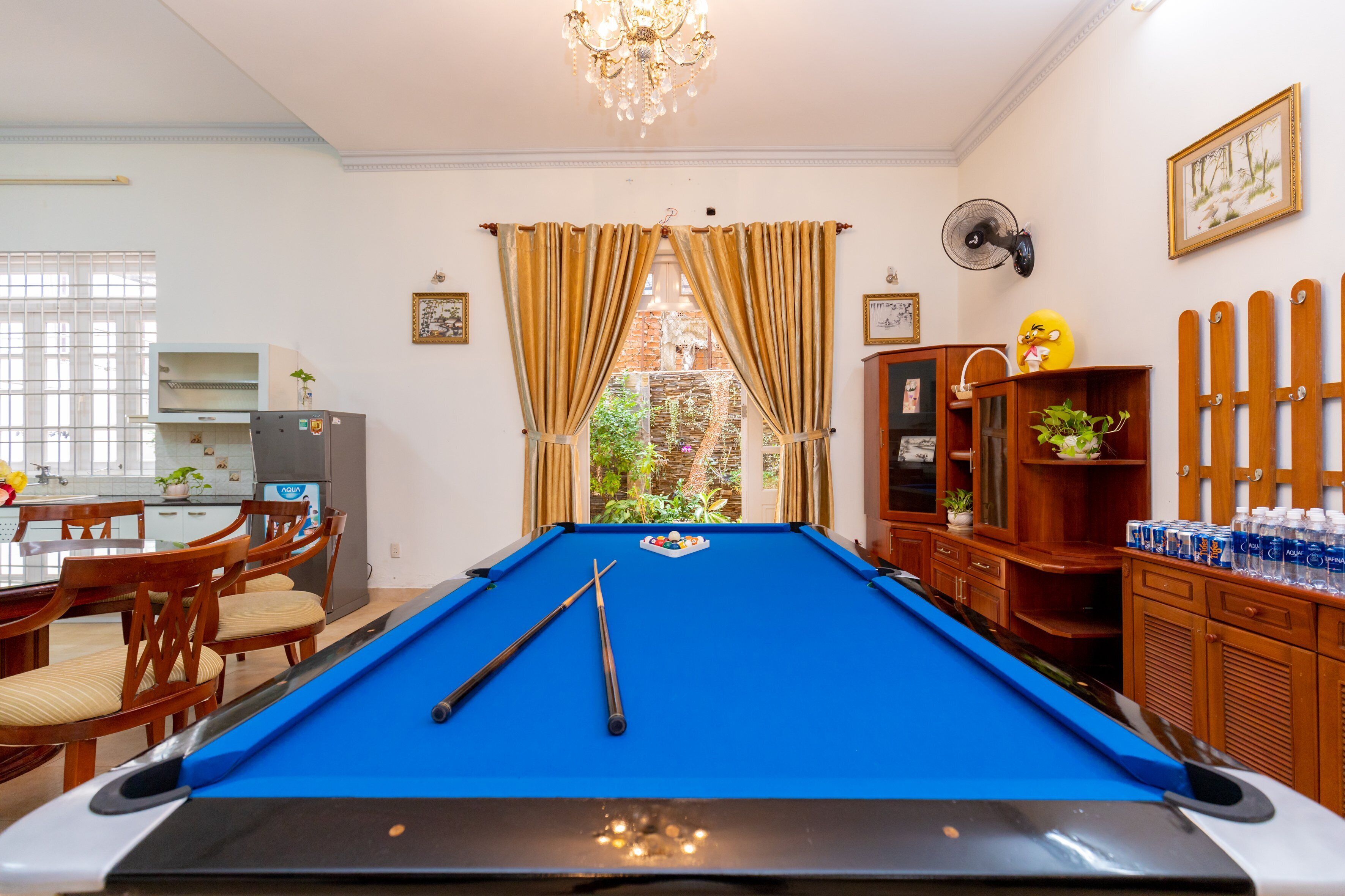 Ein Billardtisch mit zwei Spielstöcken, ein Essbereich mit Holzsesseln, ein Kühlschrank und Blick ins Grüne durch ein Fenster mit Vorhängen.