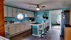 A compact kitchen with white cabinets, a central island with stools, a ceiling fan, and a refrigerator. - Bella Vista - Waterfront Getaway, Kayaks, fishing & Close to Topsail Beaches! (Sneads Ferry)