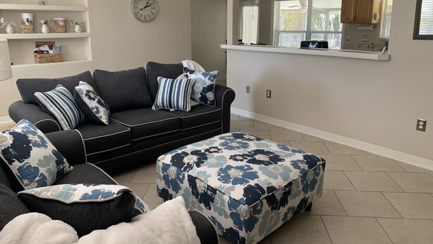 A living room with a dark grey sofa, floral ottoman, and a white clock on the wall. - Hidden gem close to everything!!! (Sarasota)
