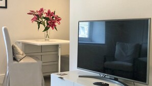 A modern living room with a flat-screen TV, a white desk, a chair, and a vase with pink flowers. - Newly refurbished, modern apartment in Hathersage, with parking (Hathersage)