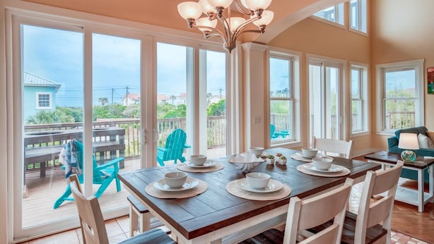 A dining area with a large table set for four, surrounded by chairs, with a view of the outdoors through large windows. - Triple Level House, Short Walk to Beach, Views, Multiple Balconies, Game Room, $300 in Free Tickets! (St. Augustine)