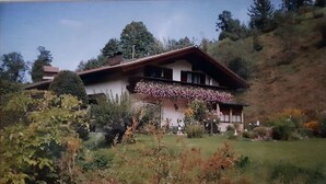 A traditional chalet with a sloped roof, surrounded by lush greenery and trees.