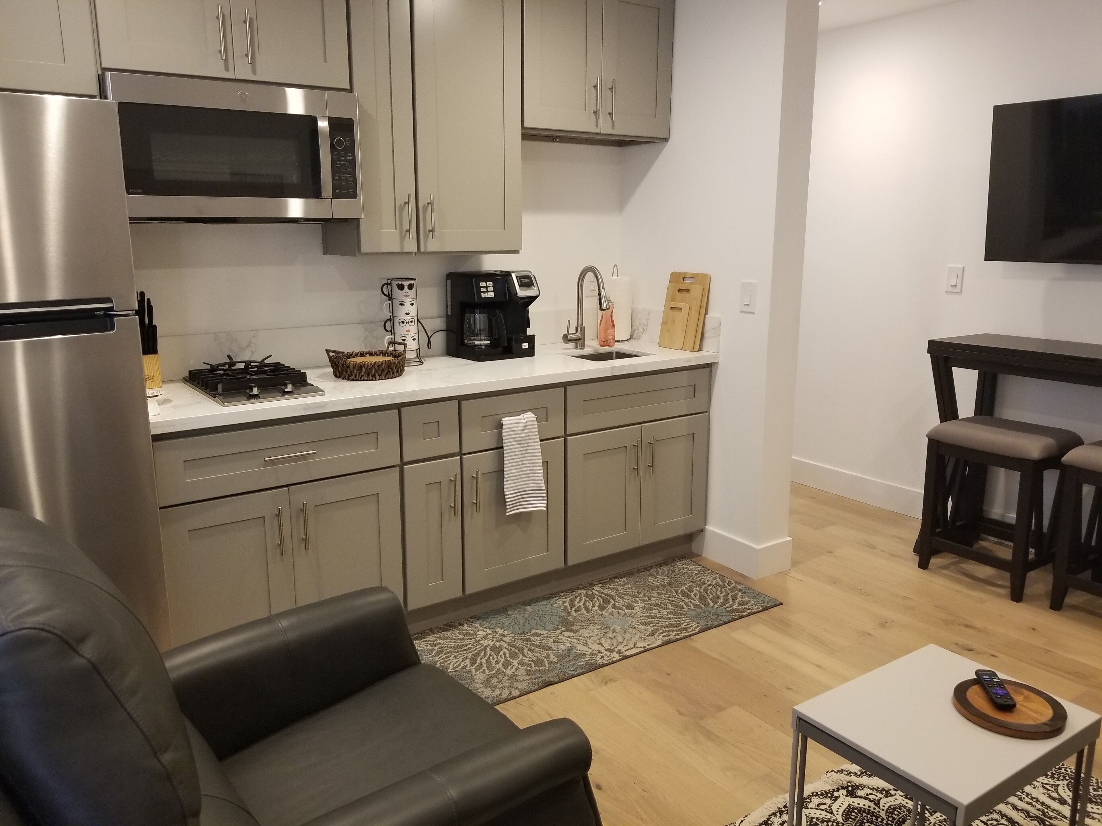 A modern kitchen with grey cabinets, a stainless steel refrigerator, a microwave, and a coffee maker. There is a small dining area with a table and two stools.