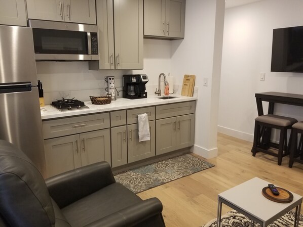 A modern kitchen with grey cabinets, a stainless steel refrigerator, a microwave, and a coffee maker. There is a small dining area with a table and two stools.