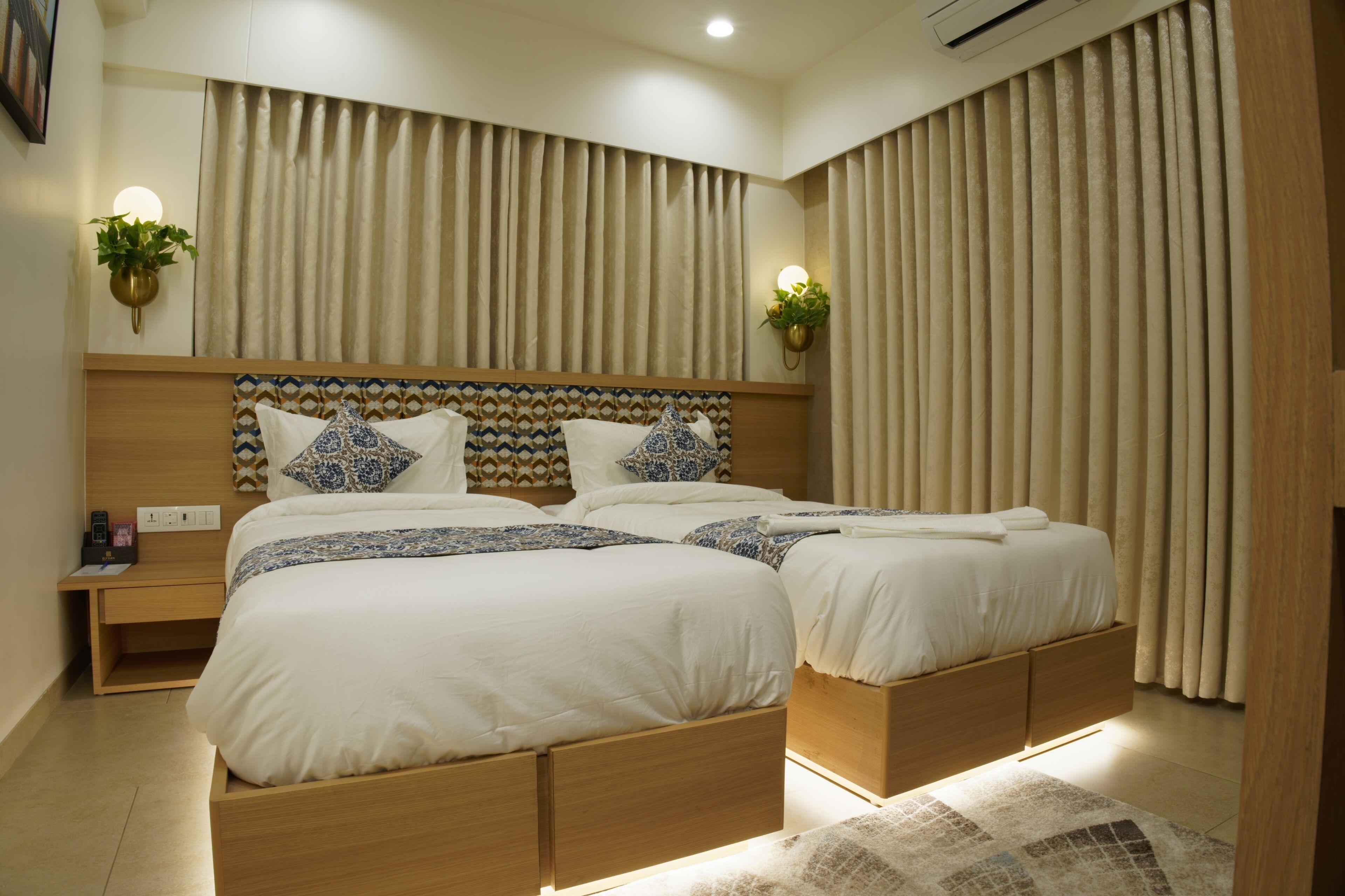 A modern bedroom with two beds, a wooden headboard, bedside tables with lamps, and a large window with curtains.