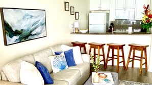 Ein Wohnzimmer mit einer Couch, einem Couchtisch und einem Teppich. Im Hintergrund ist eine Küche mit weißen Schränken und einer Arbeitsplatte zu sehen.