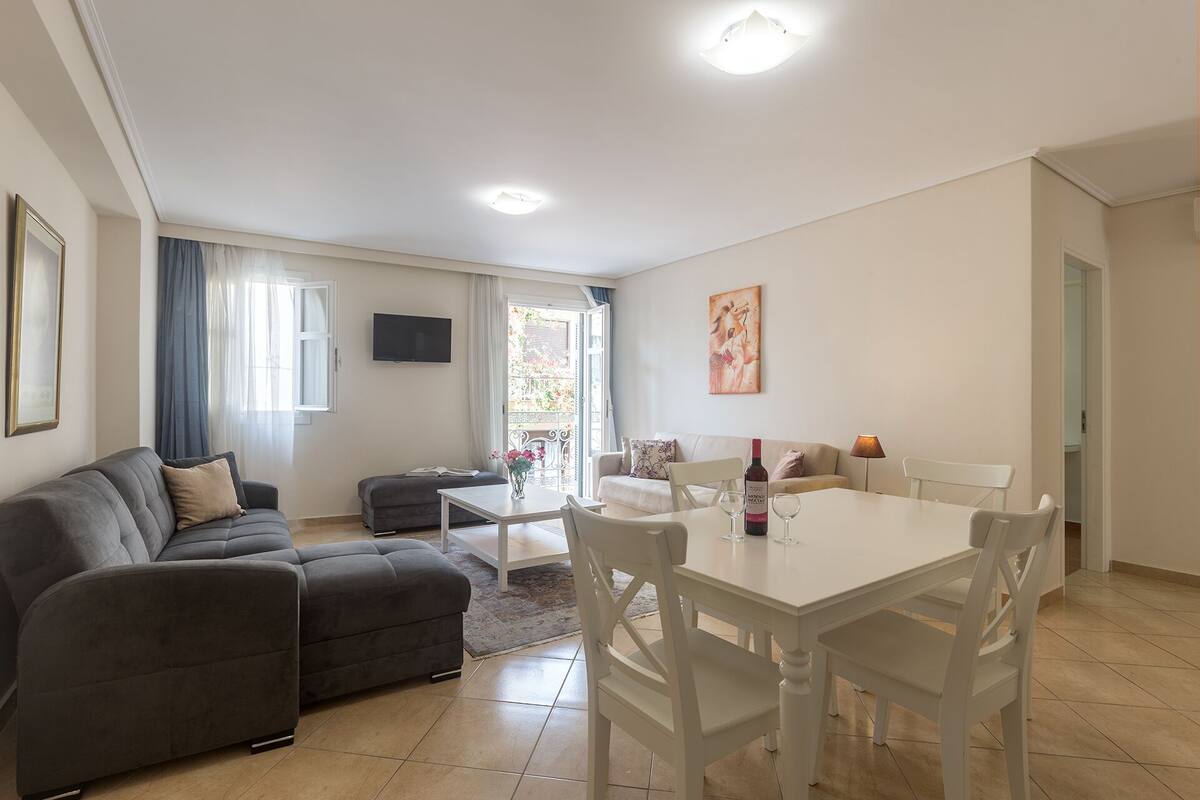 Uma sala de estar moderna com um sofá cinzento, uma mesa de jantar branca com cadeiras e uma TV fixada na parede.
