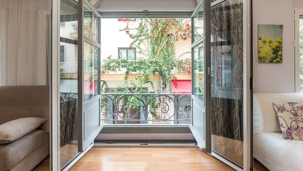 A room with a sliding glass door leading to a balcony with a view of a building and a tree with red flowers. - UrbanStay (Athens)