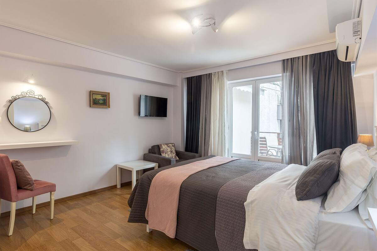 Quarto moderno com cama, escrivaninha pequena, cadeira, televisão e janela com cortinas.